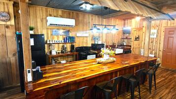 A wooden bar area with a long counter, bar stools, and a kitchen in the background.