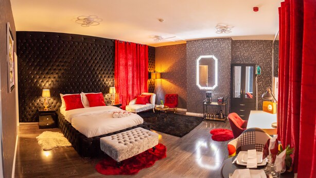 Une chambre d’hôtel moderne avec un grand lit, un coin salon comprenant un fauteuil rouge, une petite table et un miroir.