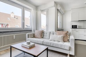 A modern apartment with a grey sofa, a small table with books, and a kitchen area. - Dreamy Iselin 2 Room w/ Storage, 1min to Local Market, by Blueground (Basel)