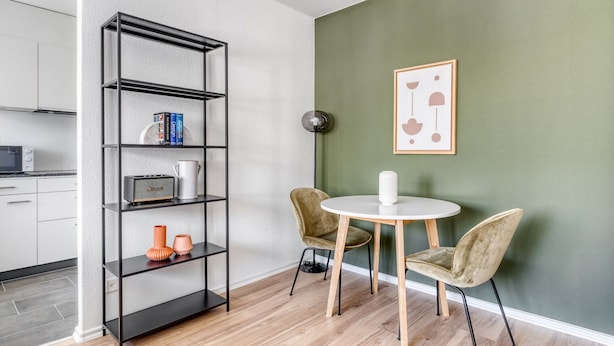 A modern living room with a black metal bookshelf, two chairs, a round white table, and a framed abstract artwork on the green wall. - Dreamy Iselin 2 Room w/ Storage, 1min to Local Market, by Blueground (Basel)