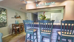 A dining area with wooden chairs and a table, a kitchen with a refrigerator, and a sign above the counter. - Oceanfront Condo w/ ocean view private balcony, on-site heated pool & hot tub (Cape Canaveral)