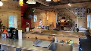 Un intérieur de chalet chaleureux, avec un revêtement mural en bois, un coin cuisine équipé d’une machine à café et un espace repas agrémenté d’un sapin de Noël.