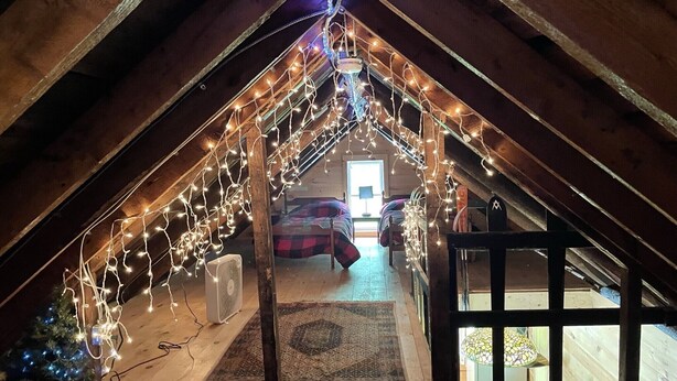 Une chambre mansardée avec un plafond en pente, des poutres en bois, un lit, un ventilateur et des guirlandes lumineuses.