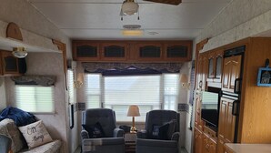 Een compact camperinterieur met twee zitbanken, een houten salontafel, een kitchenette met een kookplaat, en een plafondventilator.