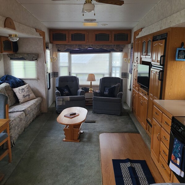 A compact RV interior with two sofas, a wooden coffee table, a kitchenette with a stove, and a ceiling fan.