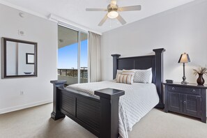 Ein Schlafzimmer mit einem großen Bett, einem Nachttisch, einer Kommode, einem Deckenventilator und einem großen Fenster mit Vorhängen.