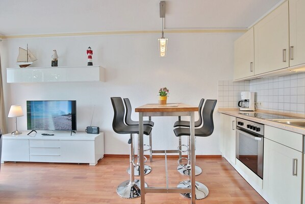 A modern kitchen with a dining area, featuring a wooden table, black chairs, and a TV on a white cabinet.