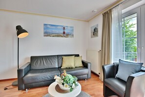 A modern living room with a grey leather sofa, a round coffee table with a floral centerpiece, a black floor lamp, and a large window with curtains.