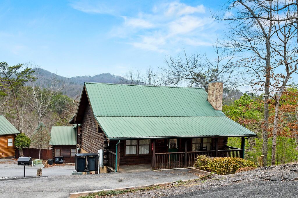 A rustic log cabin with a green roof, a porch, and a small front yard.
