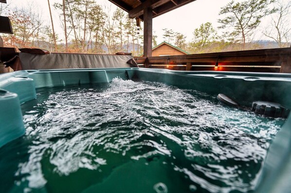 A hot tub with water splashing, surrounded by a wooden deck and a covered area with a view of trees and a building in the background. - 1BR Cozy Cabin - Romantic, Hot Tub, Couples, RELAX in Pigeon Forge (Pigeon Forge)