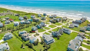 Vue aérienne d’un quartier résidentiel situé sur la côte, avec des maisons, des pelouses verdoyantes et une plage.
