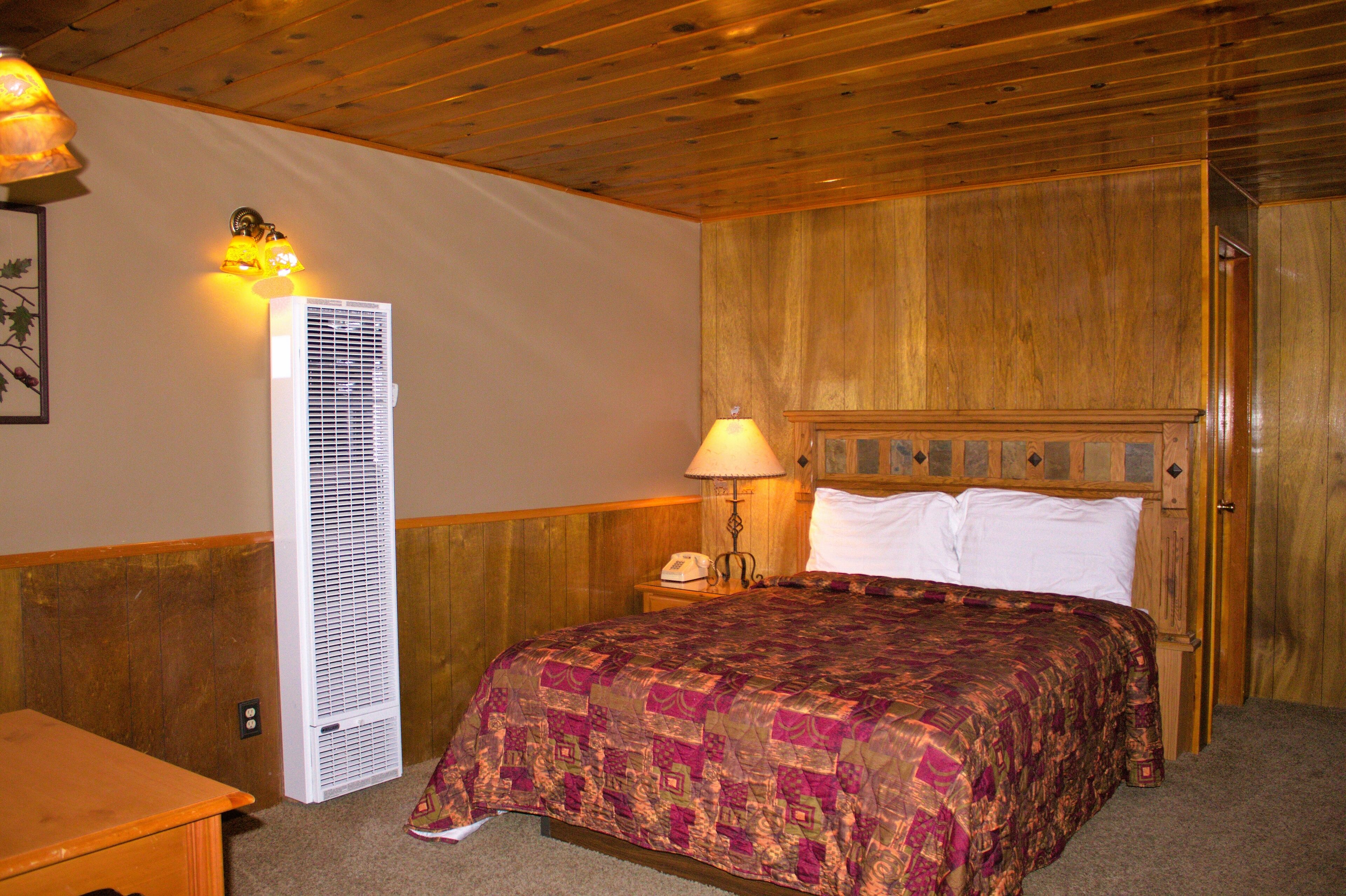 A bedroom with a wooden headboard, a bed with a patterned bedspread, a standing heater, a nightstand with a lamp, and a telephone.