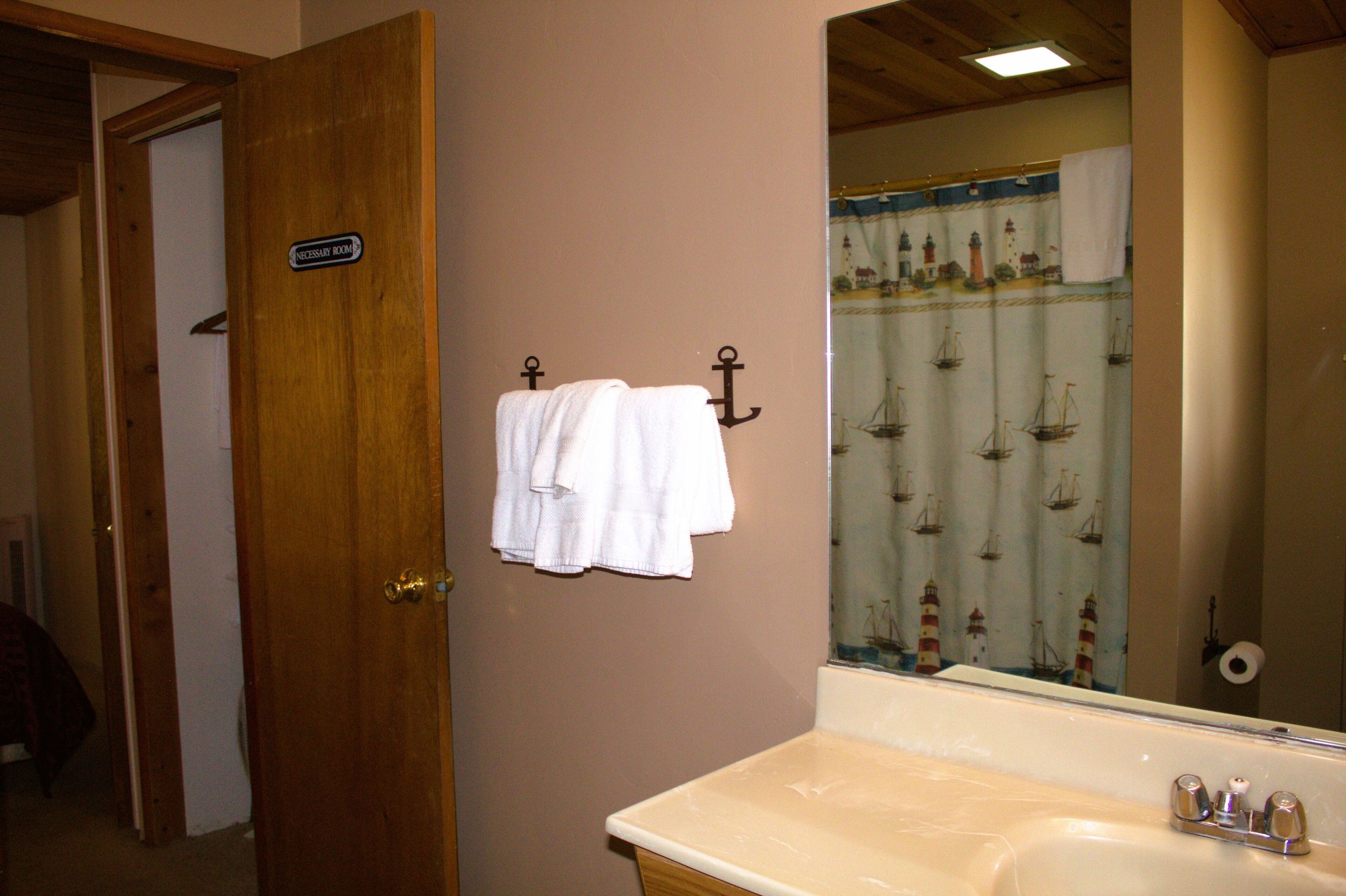Ein Badezimmer mit Holztür, einem weißen Handtuch an einem Haken, einem Spiegel, der einen Vorhang mit Meeresmotiven reflektiert, und einer Badewanne mit Wasserhahn.