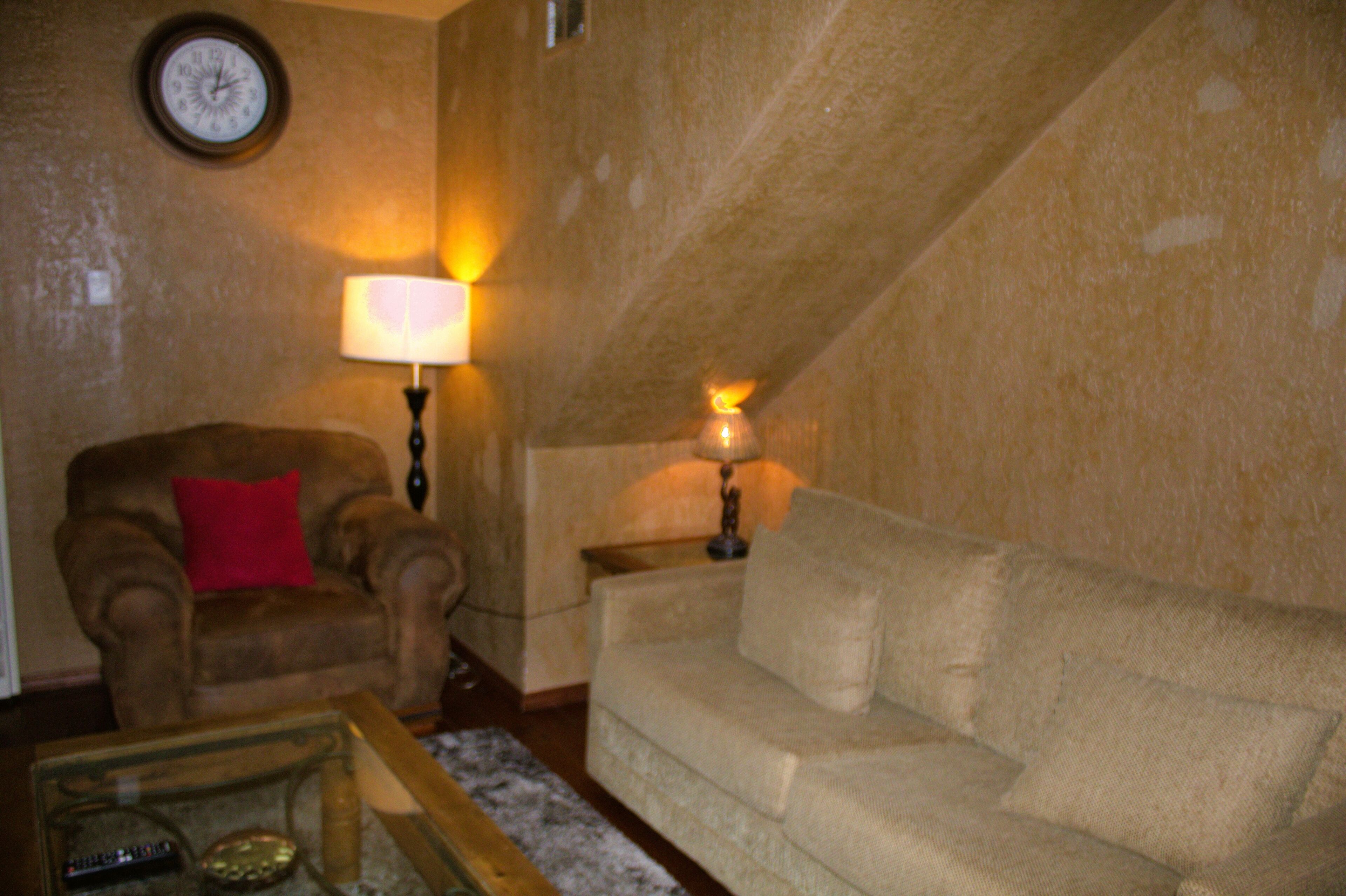 Una sala de estar con un sillón marrón, un sofá beige, una mesa de centro de vidrio y dos lámparas de pared.