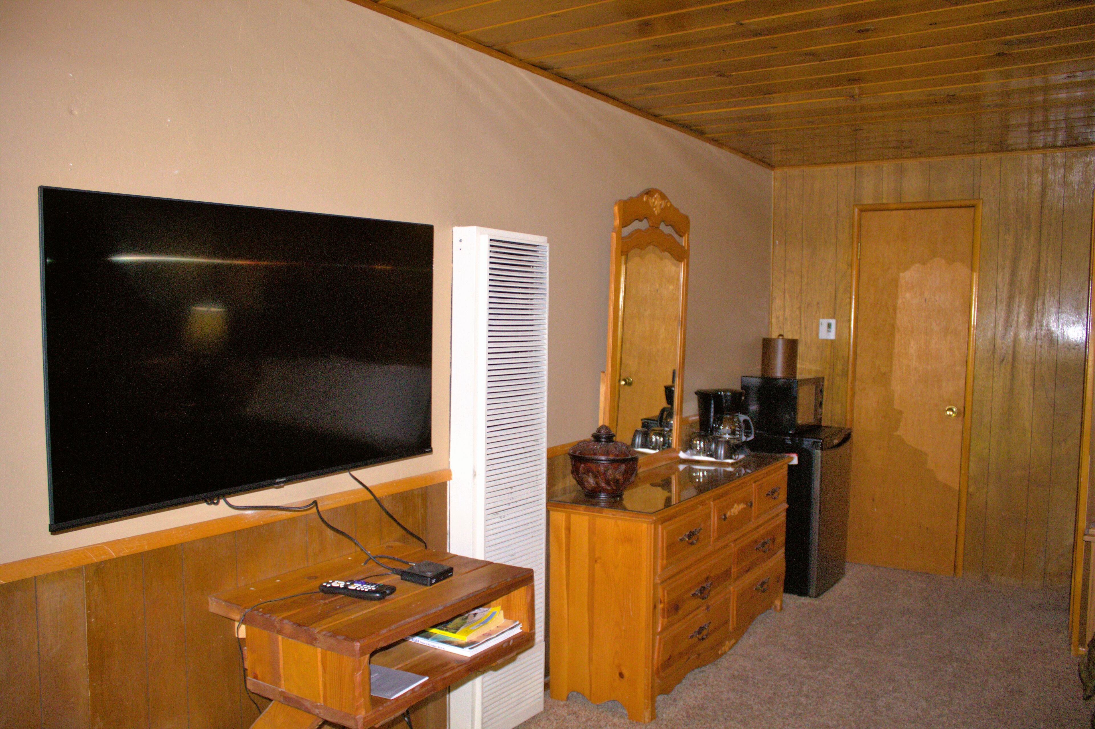 Quarto com televisão de tela plana na parede, cômoda de madeira com gavetas, espelho e cafeteira.