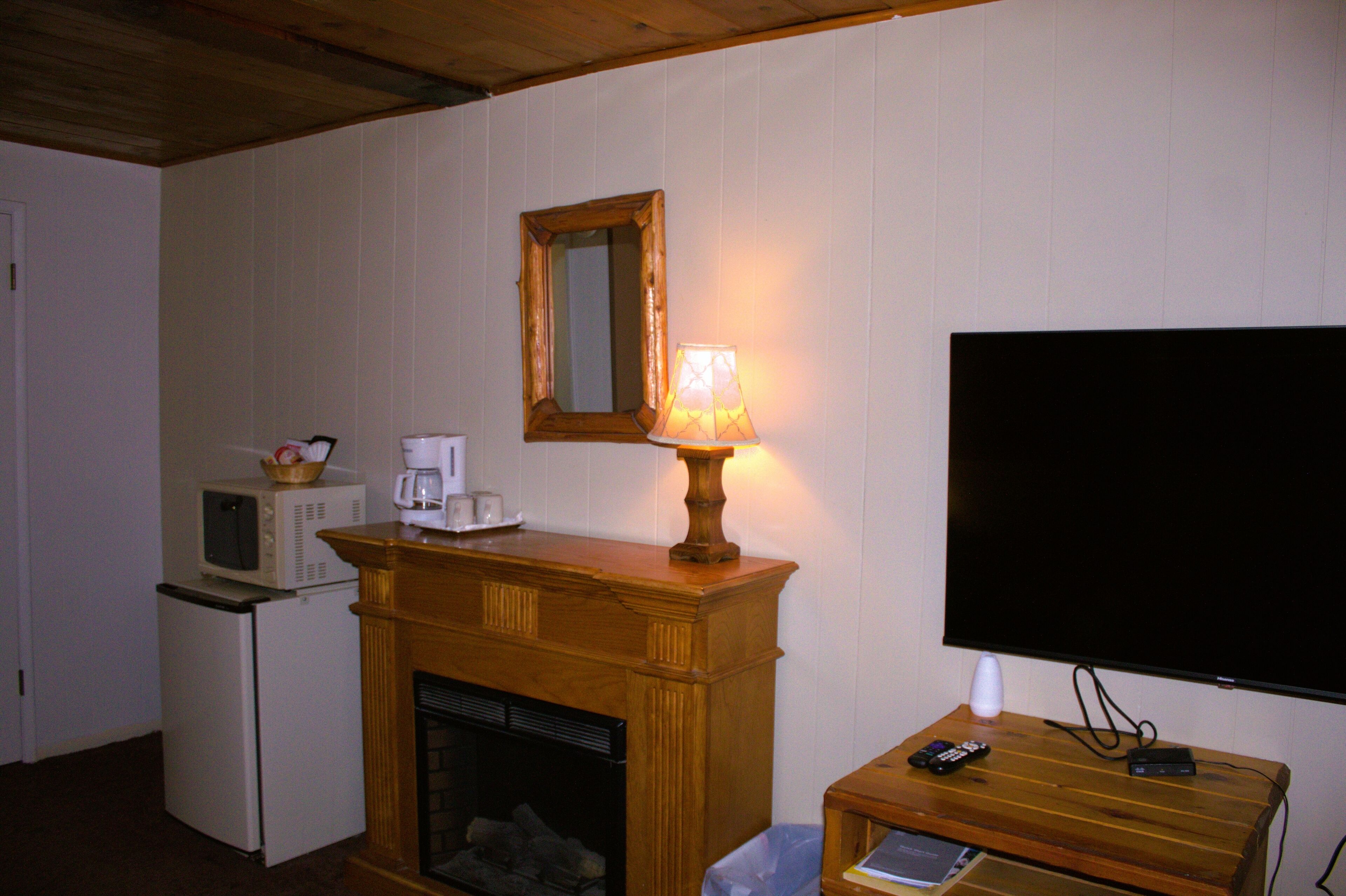 A room with a fireplace, a television mounted on the wall, a microwave, and a small table with a lamp.