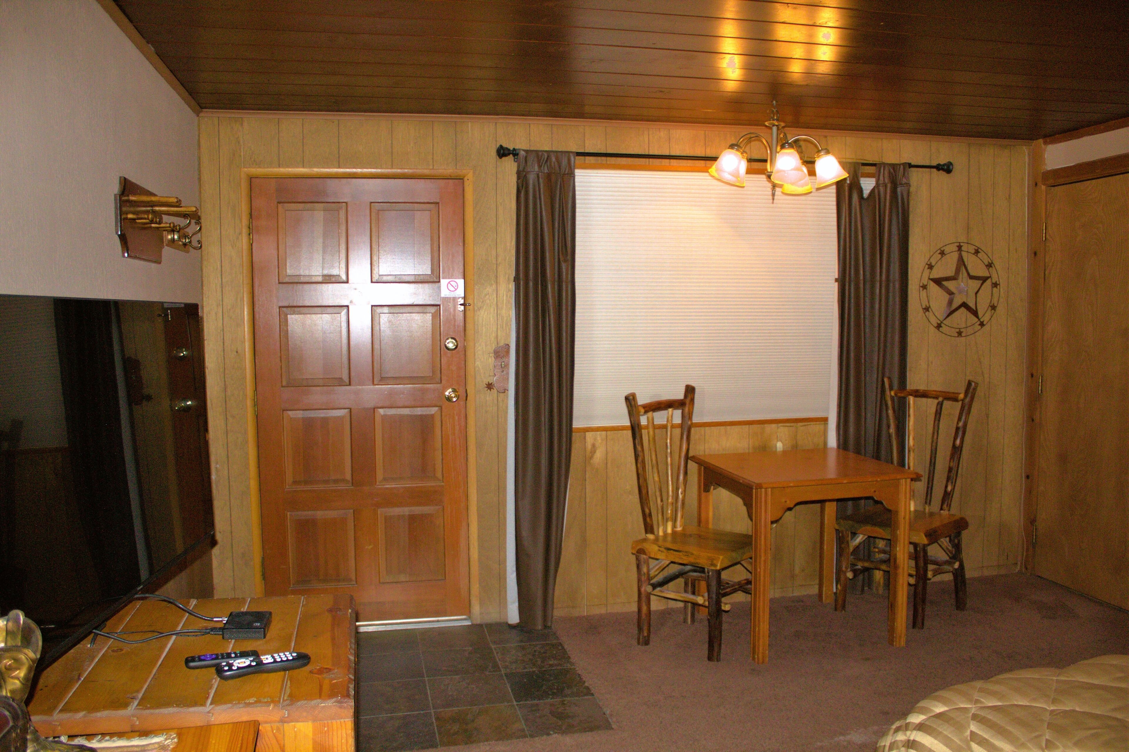 A room with a wooden door, a television, a table with chairs, and a ceiling light fixture.