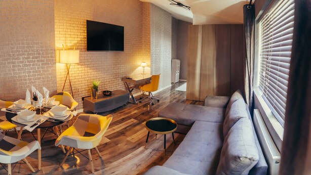 A modern living room with a brick wall, a flat-screen TV, a dining area with checkered chairs, a sofa, and a small round table. - Impeccable 1-Bed Apartment in Birmingham (Birmingham)