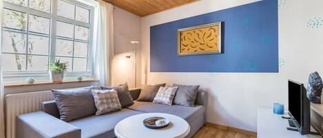 A modern living room with a grey sofa, a small round table, a blue rug, a framed artwork on the wall, and a window with a view of trees.