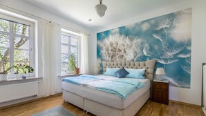 Een slaapkamer met een groot bed, een raam met gordijnen, een radiator, een wanddecoratie en een nachtkastje met een lamp.