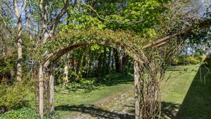 Een tuin met een houten pergola begroeid met klimplanten, een grindpad en omliggend groen.