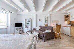 A modern, well-lit living room with a sofa, coffee table, and a television. There is a kitchen area with a dining table and chairs.