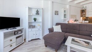 Una sala de estar con un televisor de pantalla plana, un sofá marrón, un mueble blanco con estanterías y una zona de cocina con cafetera.