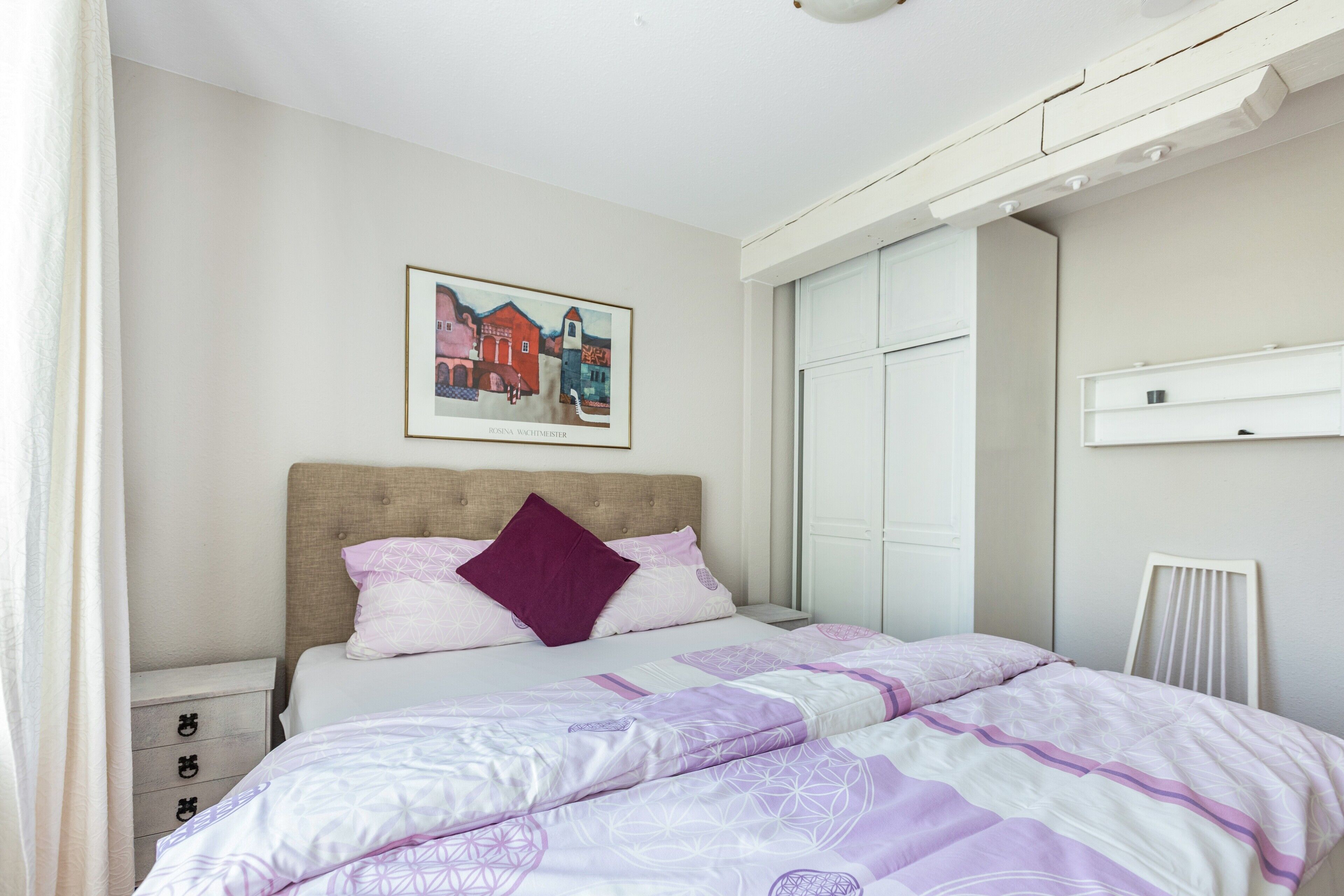 Ein Schlafzimmer mit Bett, Kopfteil, Wandbild, Kleiderschrank und Heizkörper.