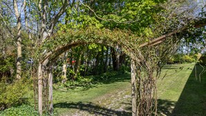 Un jardín con una pérgola de madera cubierta de hiedra, rodeado de árboles y un sendero cubierto de césped.