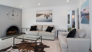 A modern living room with a fireplace, two sofas, a glass coffee table, and a large painting of a marina.