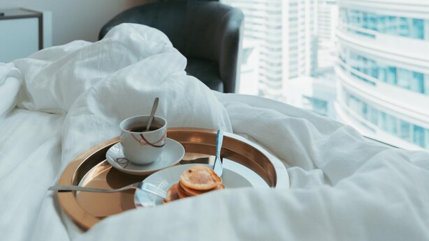 A hotel room with a bed, a tray with breakfast items, a chair, and a view of tall buildings through a window.