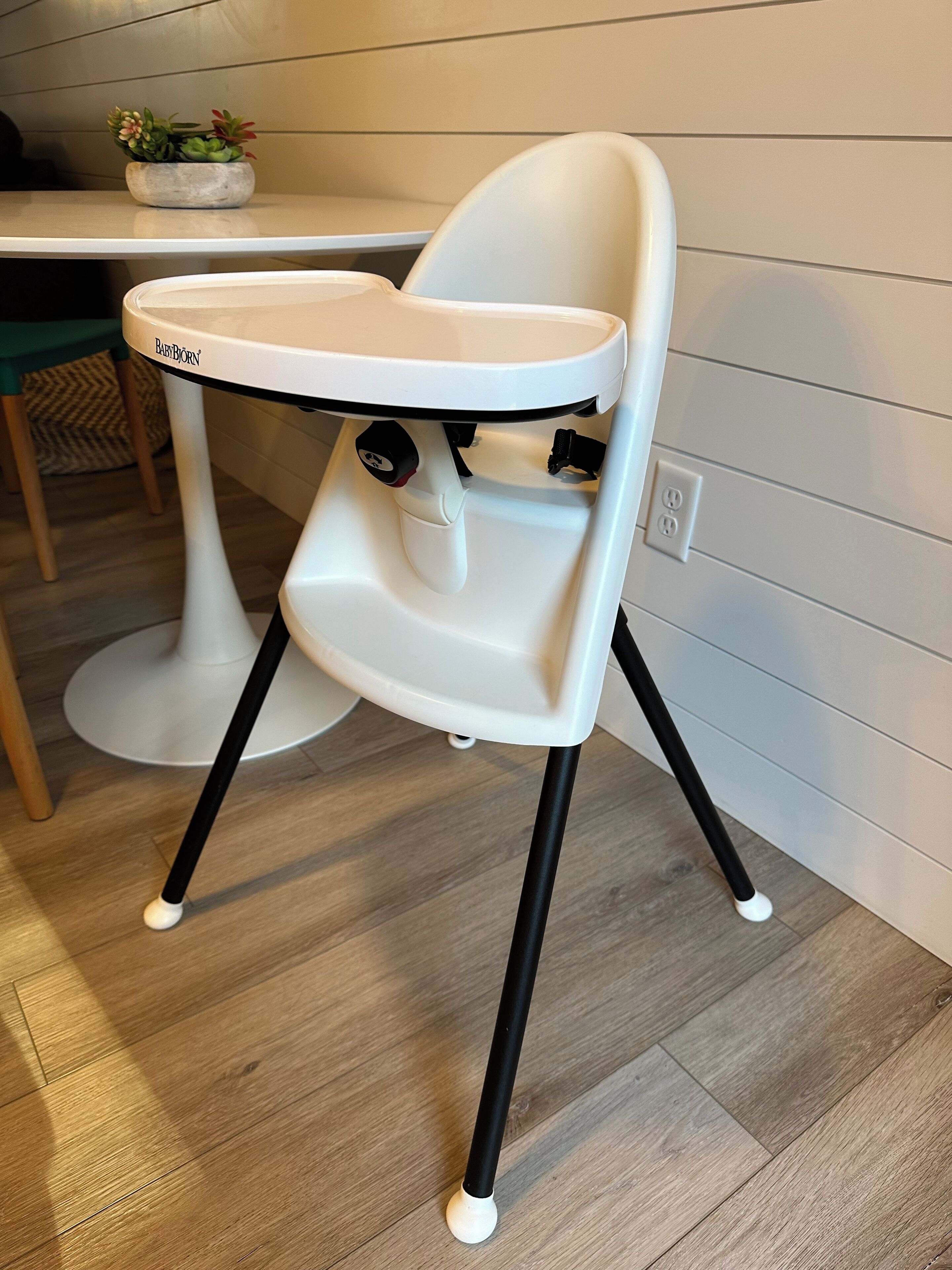 A high chair with a tray, positioned on a wooden floor with a table and chair in the background.
