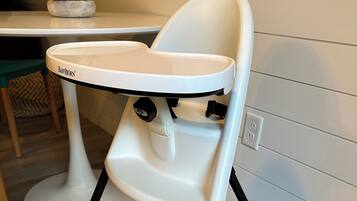 A high chair with a tray, positioned on a wooden floor with a table and chair in the background.