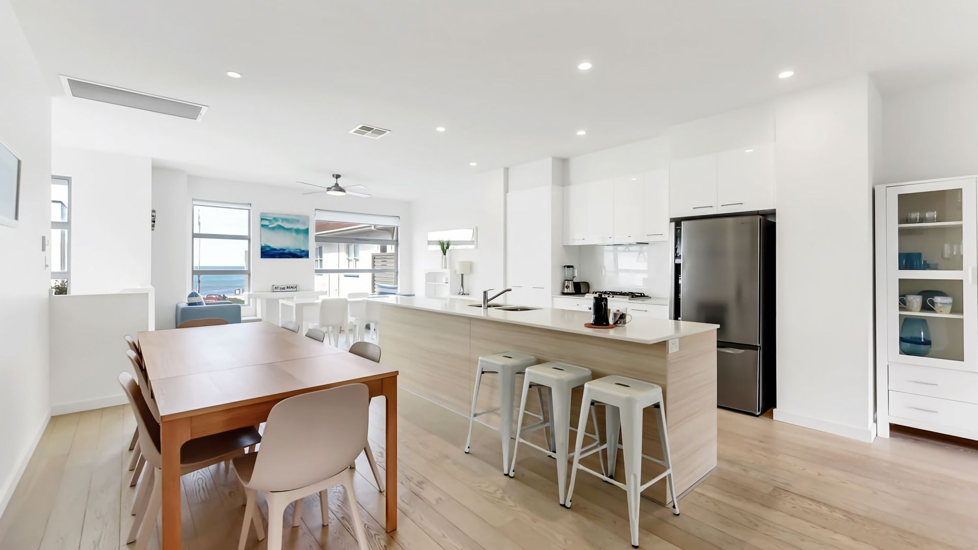 A modern kitchen with white cabinets, a stainless steel refrigerator, and a dining area with a wooden table and white chairs.