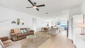 A modern living room with a sofa, coffee table, and a ceiling fan. There is a kitchen area with a dining table and chairs visible through an open door. - Stunning Sea Views in Excellent Location (Victor Harbor)