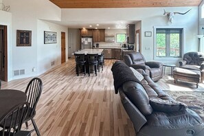 A spacious living area with a large leather sofa, a dining table with chairs, a kitchen area with wooden cabinets, and a large window with a view.