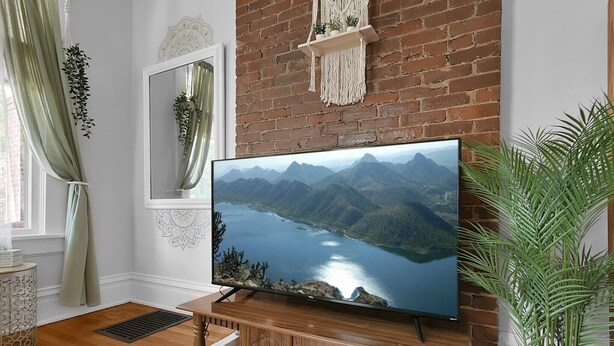 A living room with a flat-screen TV on a wooden stand, a brick wall, and a large window with curtains. - Modern Boho🪴Style 1BR Near DT on Historic Street (Columbus)