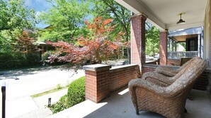A covered porch with a wicker chair, brick walls, and a view of a garden with trees. - Modern Boho🪴Style 1BR Near DT on Historic Street (Columbus)
