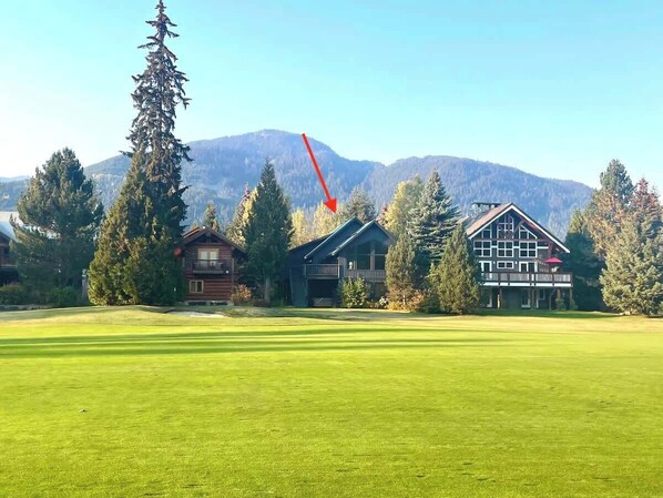 A large, open grassy area with two wooden buildings, one with a balcony and another with a red roof, surrounded by trees and mountains in the background.
