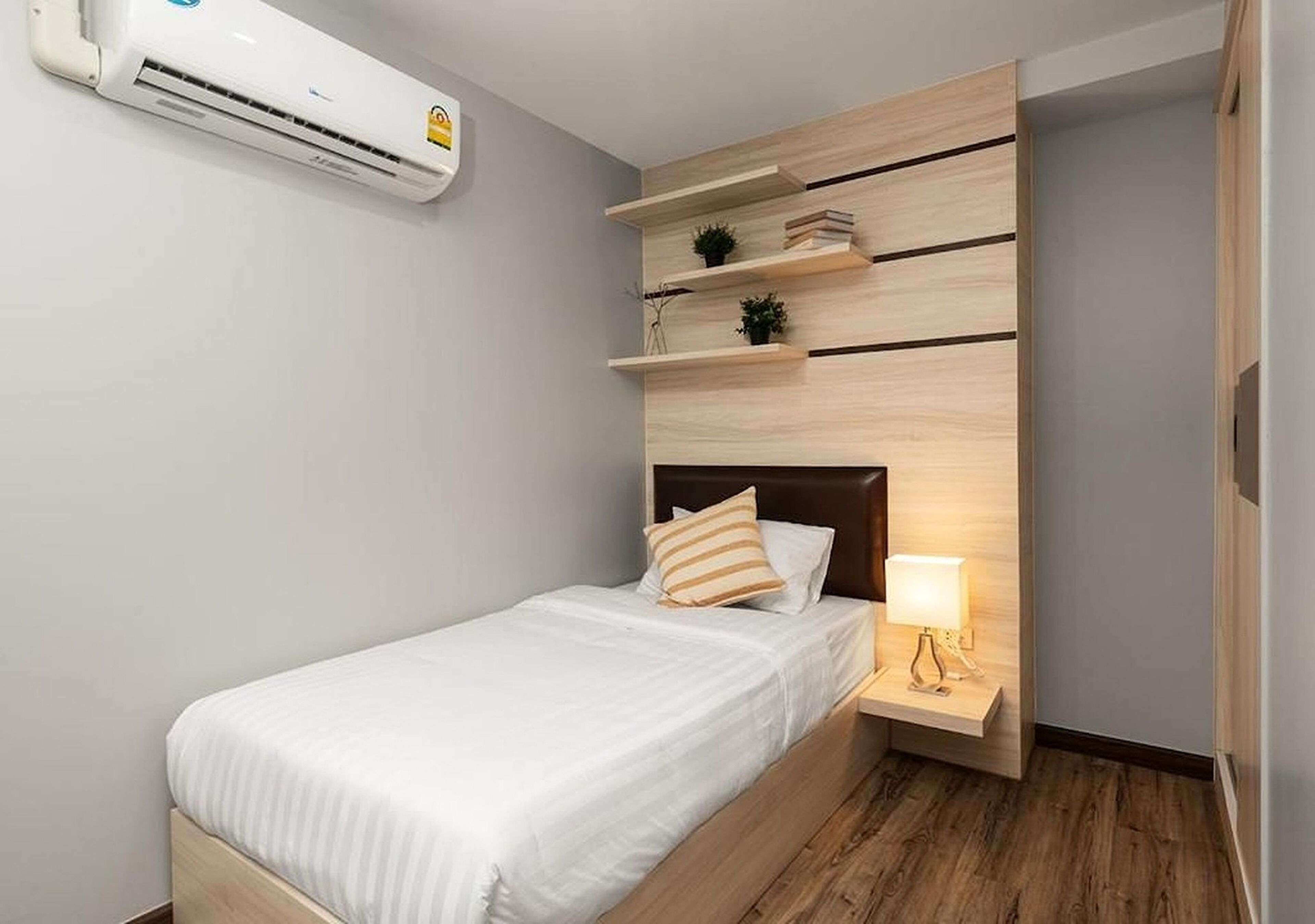 A single bed with a wooden headboard, a bedside table with a lamp, and a wall-mounted air conditioner.