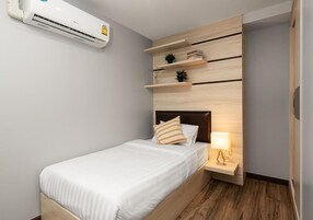 Una cama individual con cabecera de madera, una mesita de noche con lámpara, y un aire acondicionado instalado en la pared.