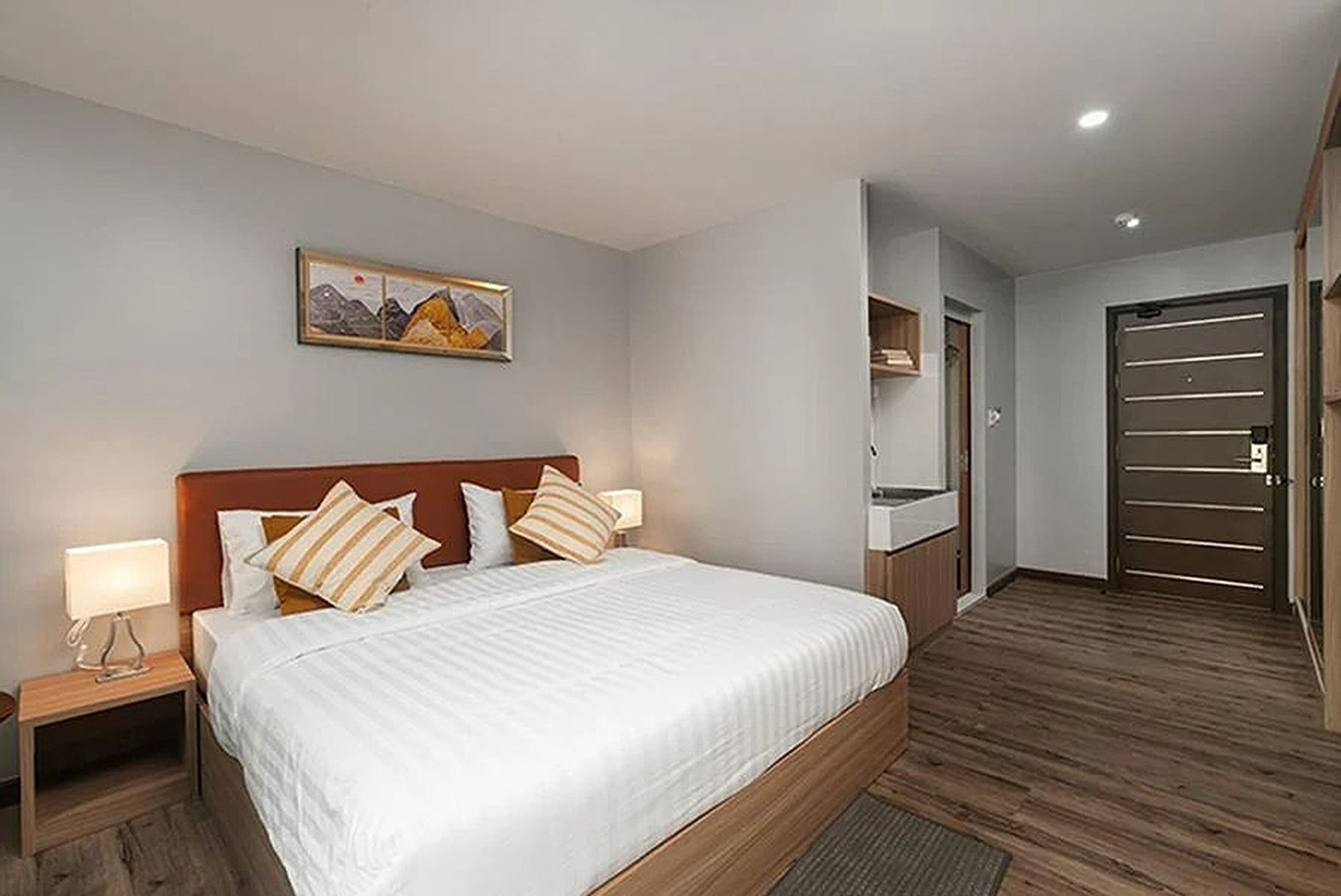 A modern hotel room with a large bed, wooden flooring, a painting on the wall, and a door with horizontal handles.