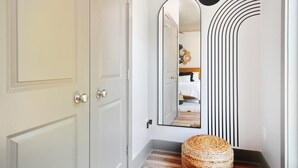A hallway with a white door, a mirror reflecting a bedroom, and a woven ottoman.