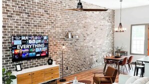 A modern living room with a brick wall, a large flat-screen TV, a sofa, a coffee table, and a dining area.