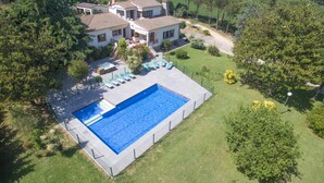 Una piscina rectangular gran amb un solàrium al voltant, a prop d'una casa de diversos pisos amb teulada enrajolada, envoltada de vegetació i arbres frondosos.