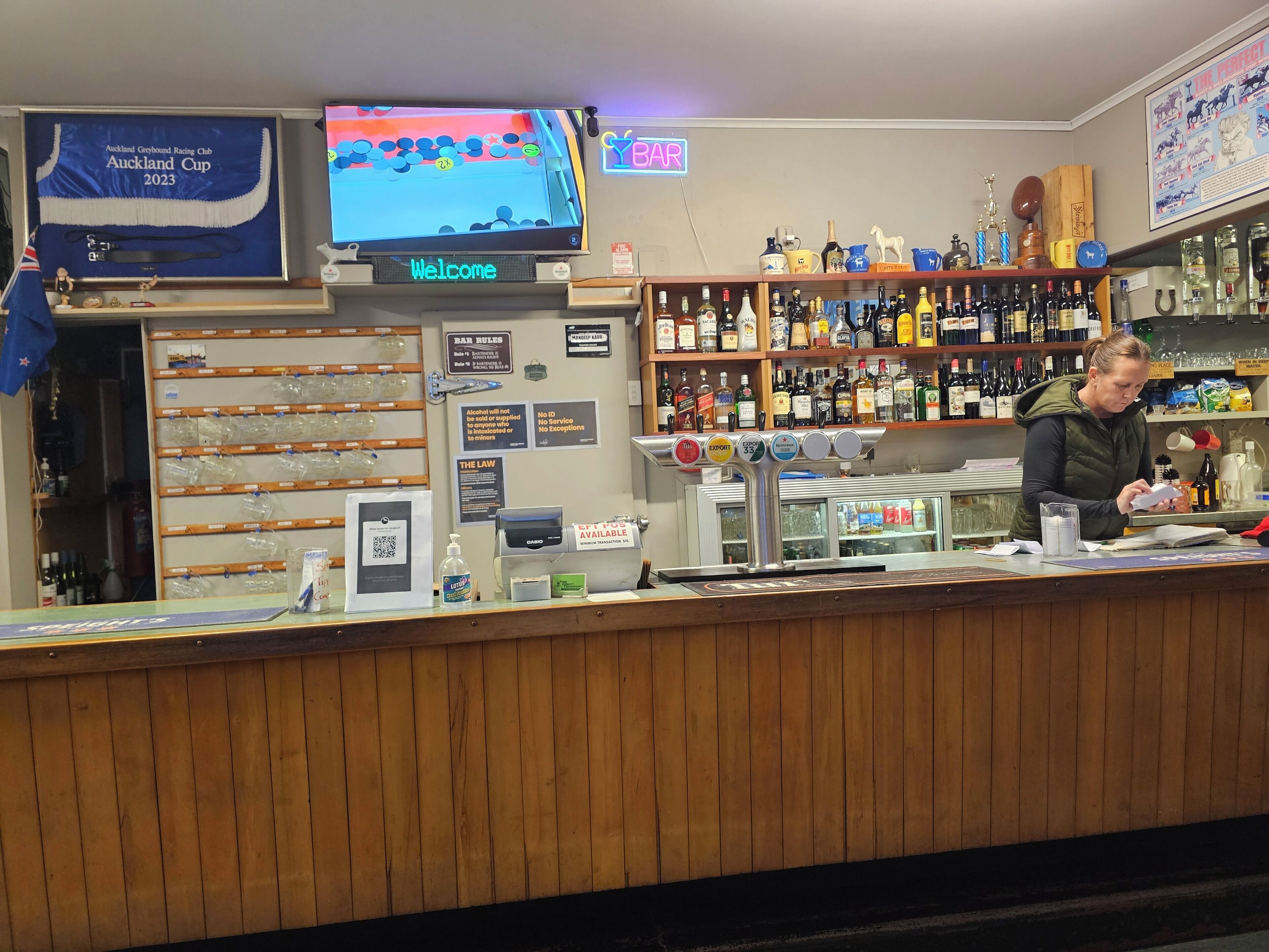 A bar area with a television displaying the Auckland Cup 2023, a bar counter with a digital 'Welcome' sign, and shelves stocked with various bottles and a 'BAR' neon sign.