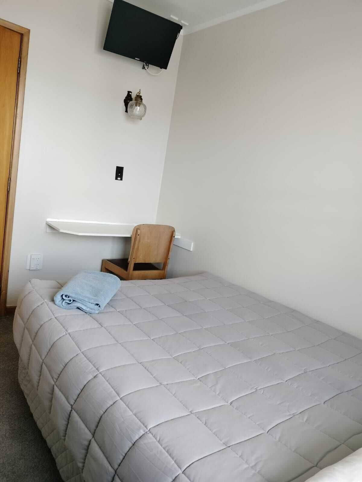 Une chambre à coucher avec un lit, une chaise en bois, un téléviseur fixé au mur et une étagère murale.