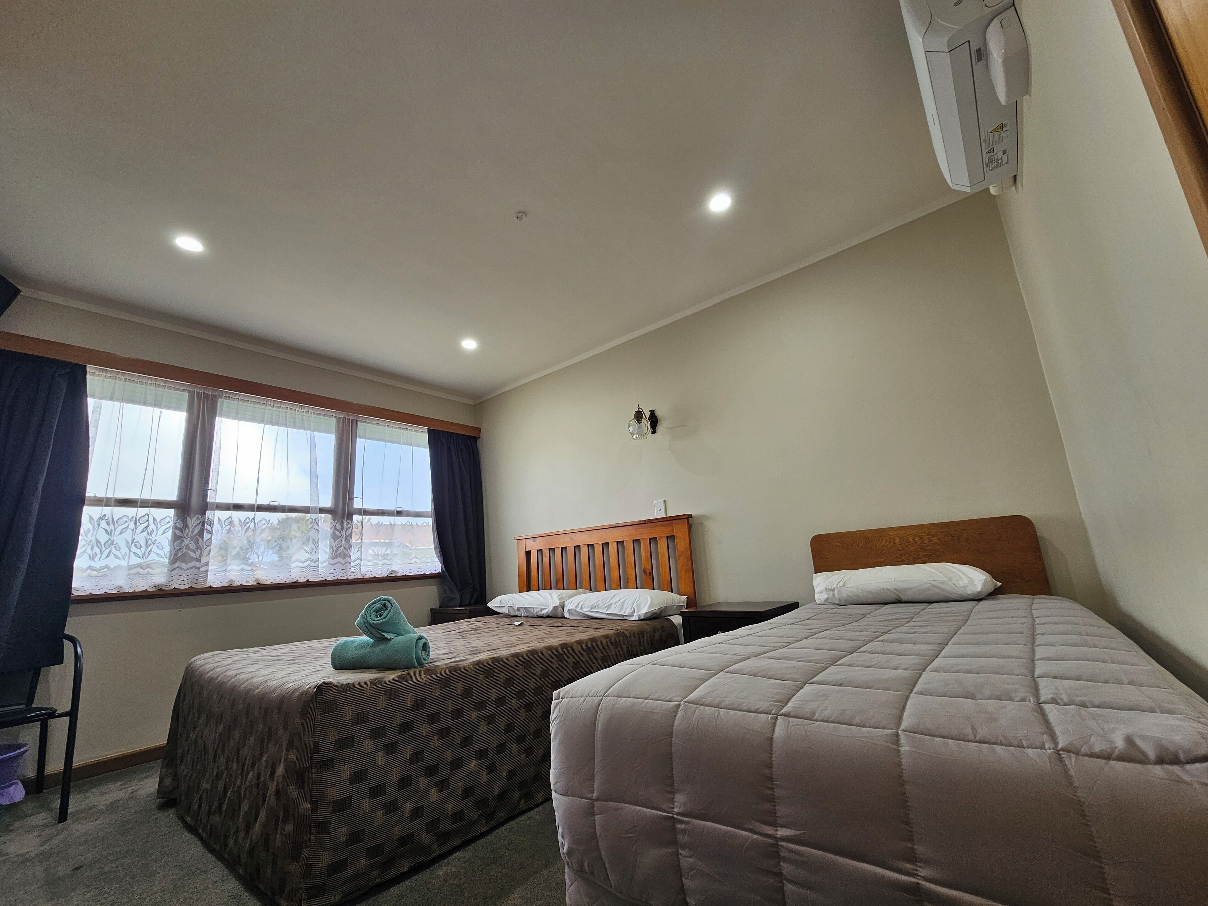 Hotellihuone, jossa on kaksi erillistä sänkyä, ikkuna verhoilla, seinään asennettu ilmastointilaite ja kattovalaisin.
