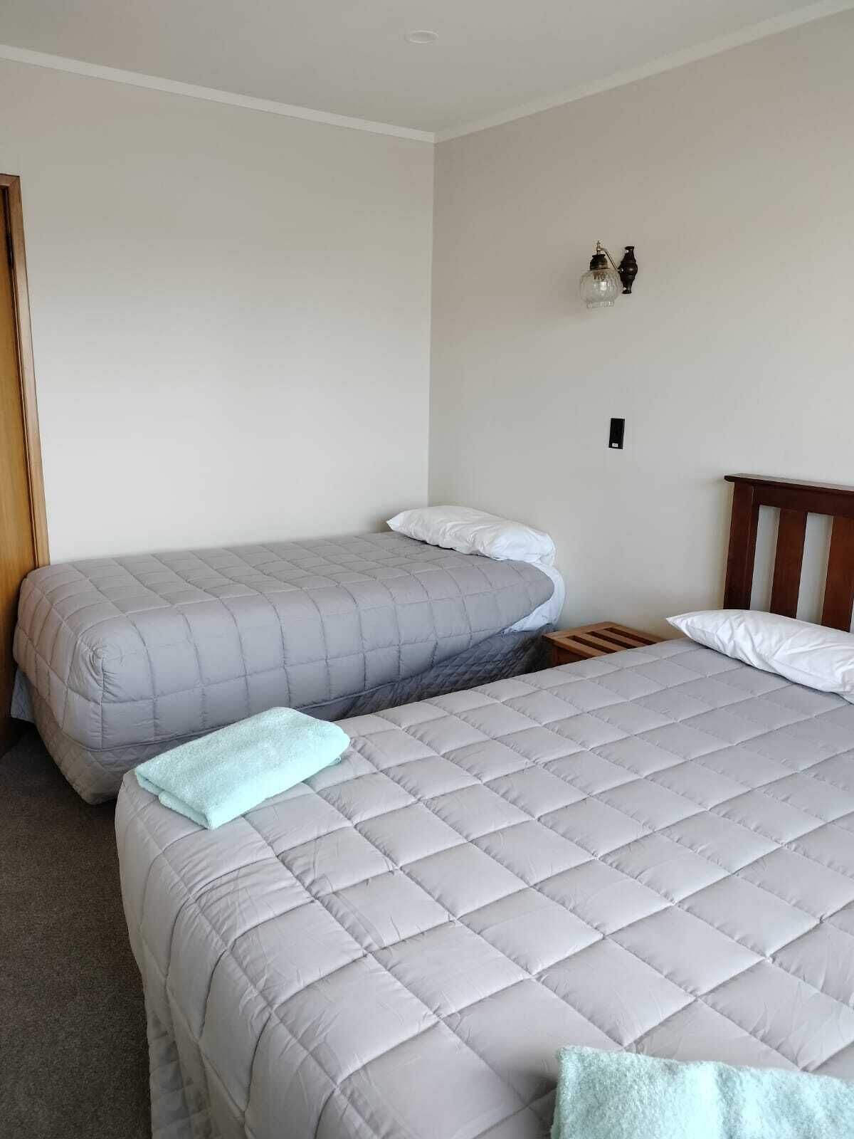 A room with two beds, each with a grey quilted mattress and white pillows. There is a wooden headboard on one bed and a small wooden nightstand with a lamp on the other.