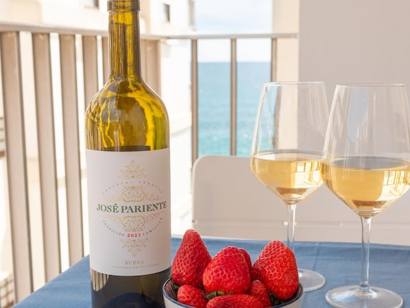 A bottle of Jose Parente wine, two glasses of white wine, and a bowl of strawberries on a table with a view of the sea.